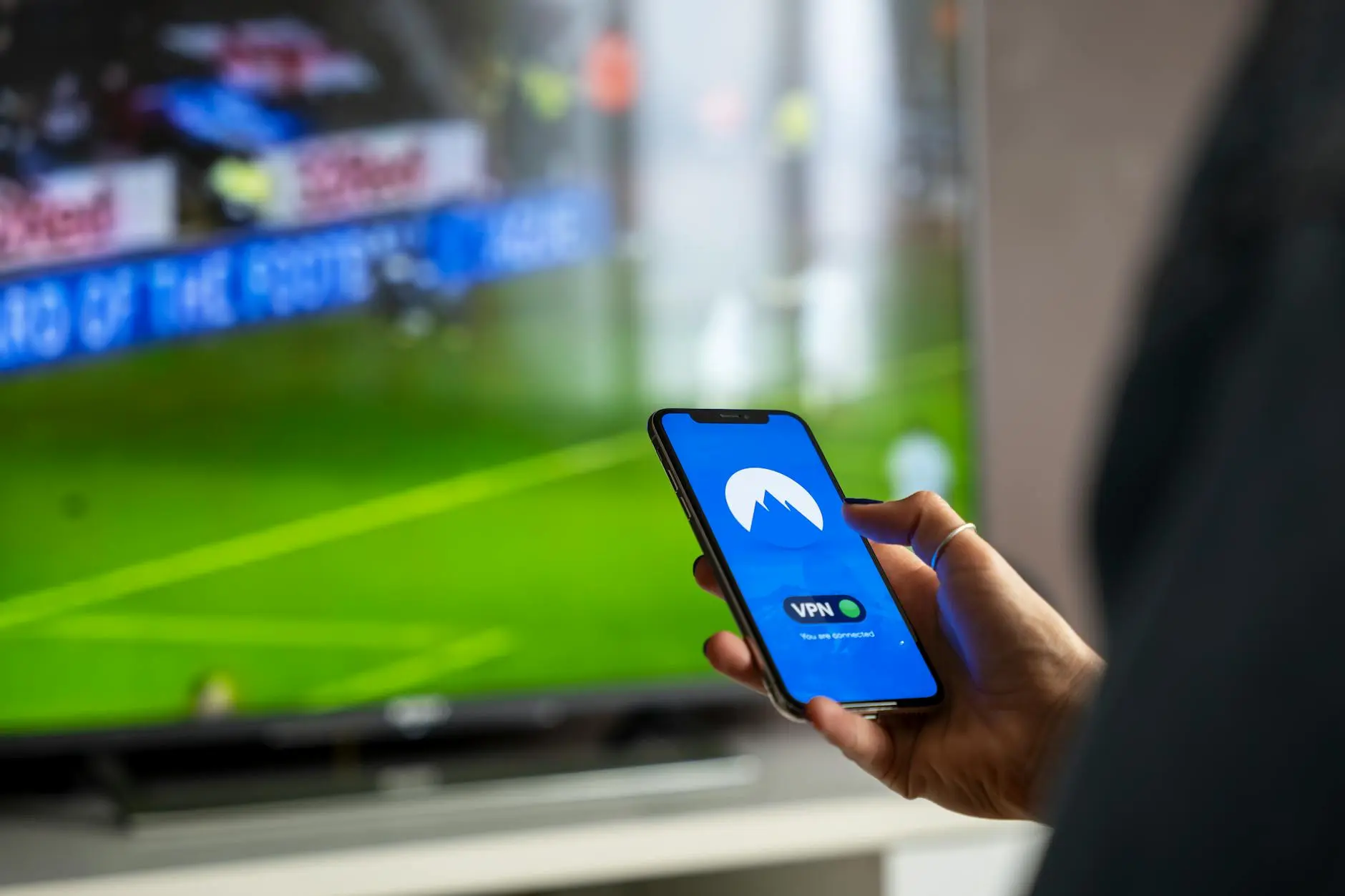 Person holding a smartphone displaying a VPN app while watching a sports event on TV.