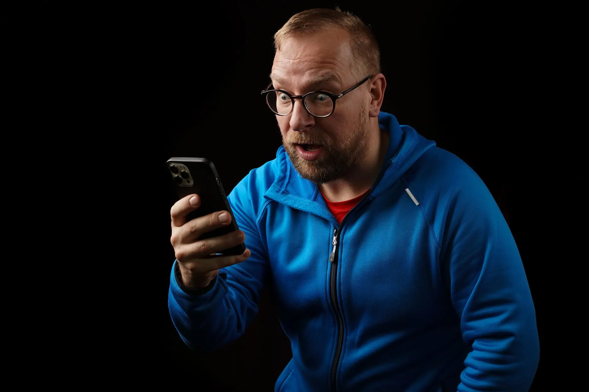 A man in a blue jacket looking surprised at his smartphone in low light.