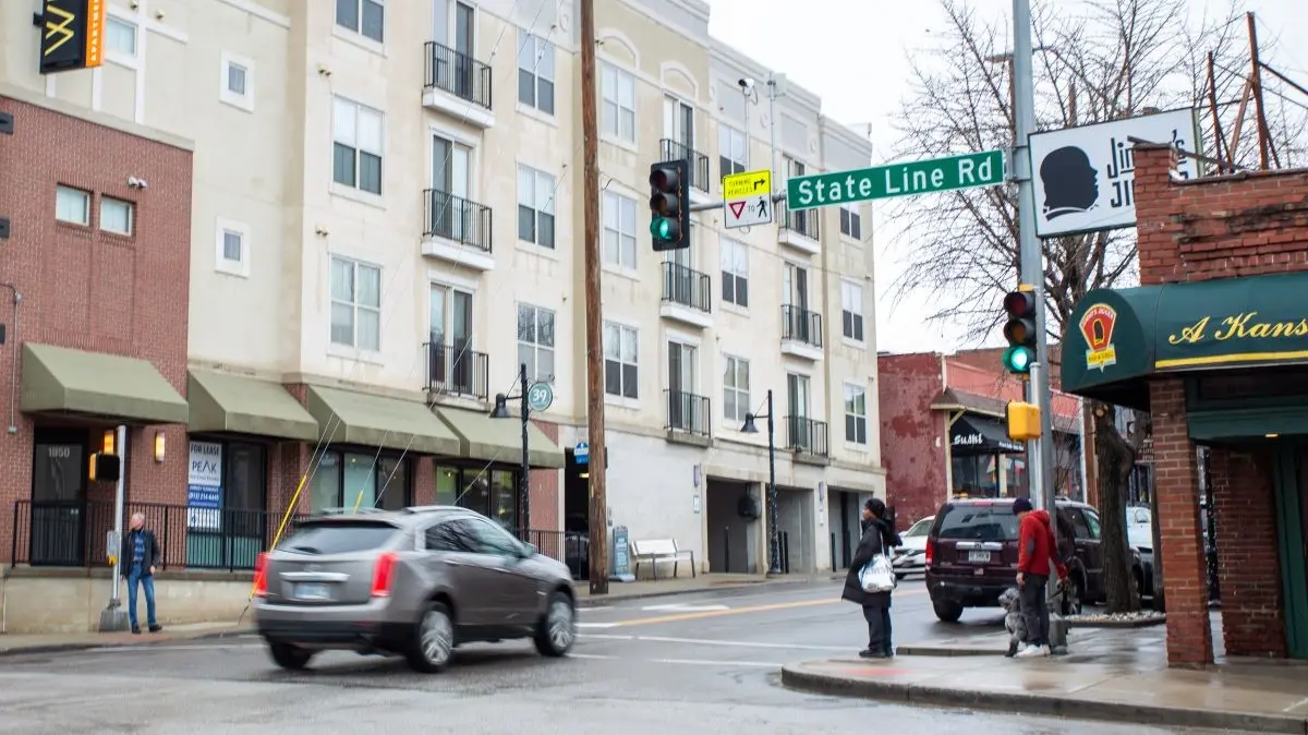 Street view of State Line Rd intersection with a green street sign, urban buildings, and passing cars.