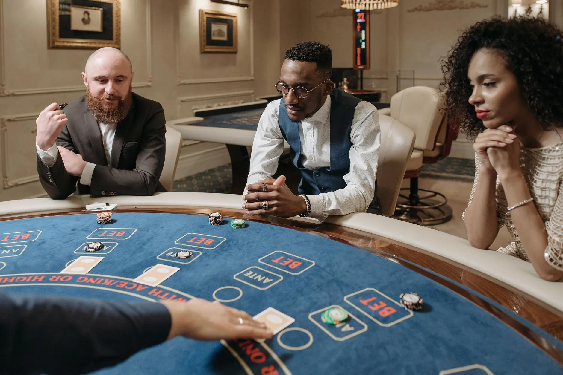 Three players at a casino baccarat table with chips and cards visible.