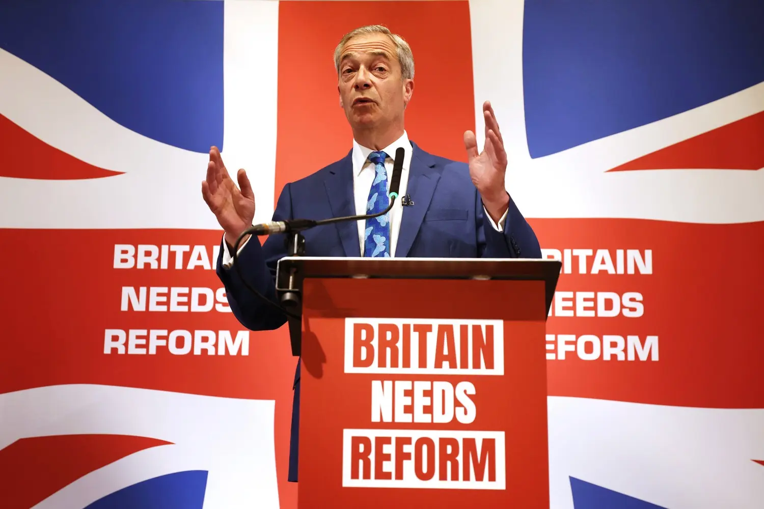 Nigel Farage speaking at a podium in front of a Union Jack background and Reform UK signage