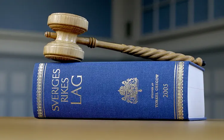 A wooden gavel rests on a blue law book titled 'Sveriges Rikes Lag'.