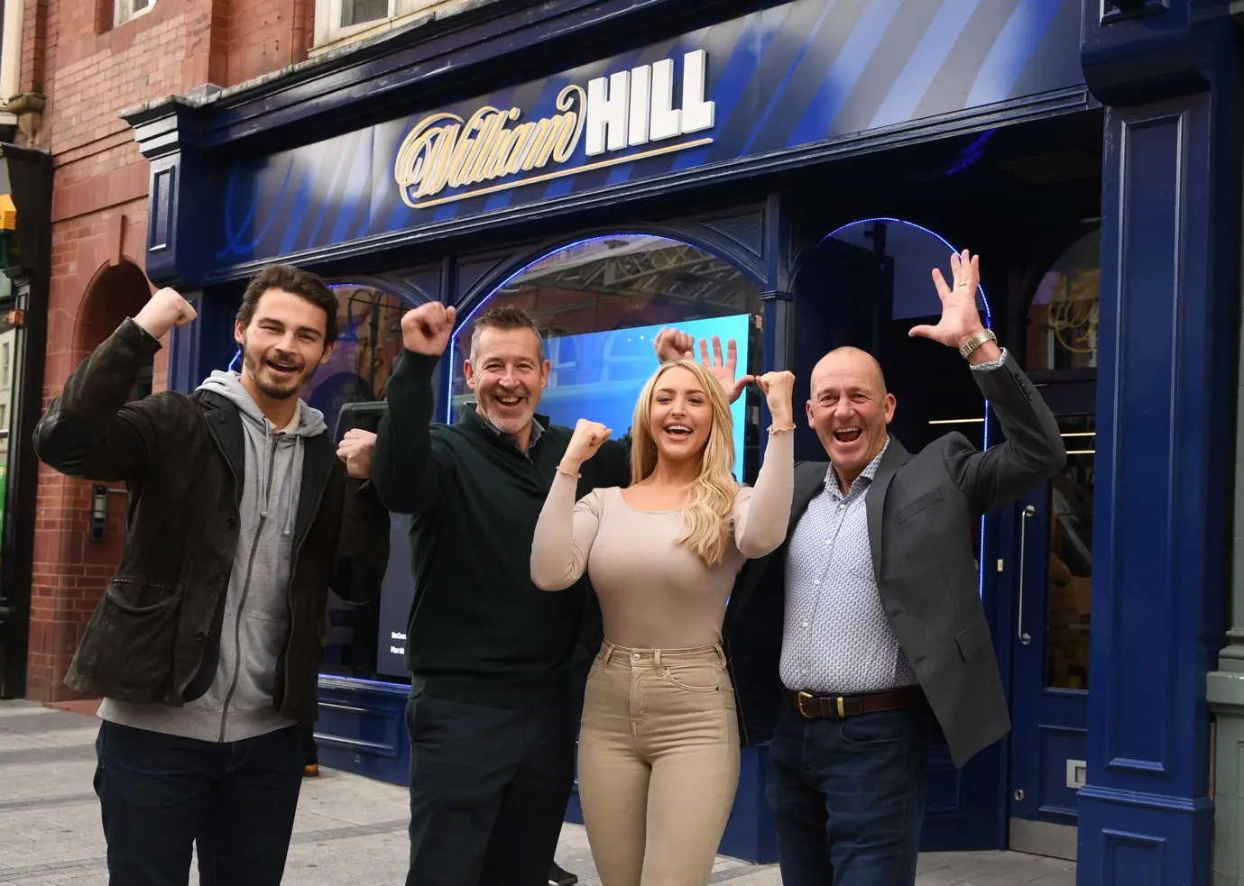 Three men and one woman cheering and celebrating in front of a blue William Hill betting shop storefront