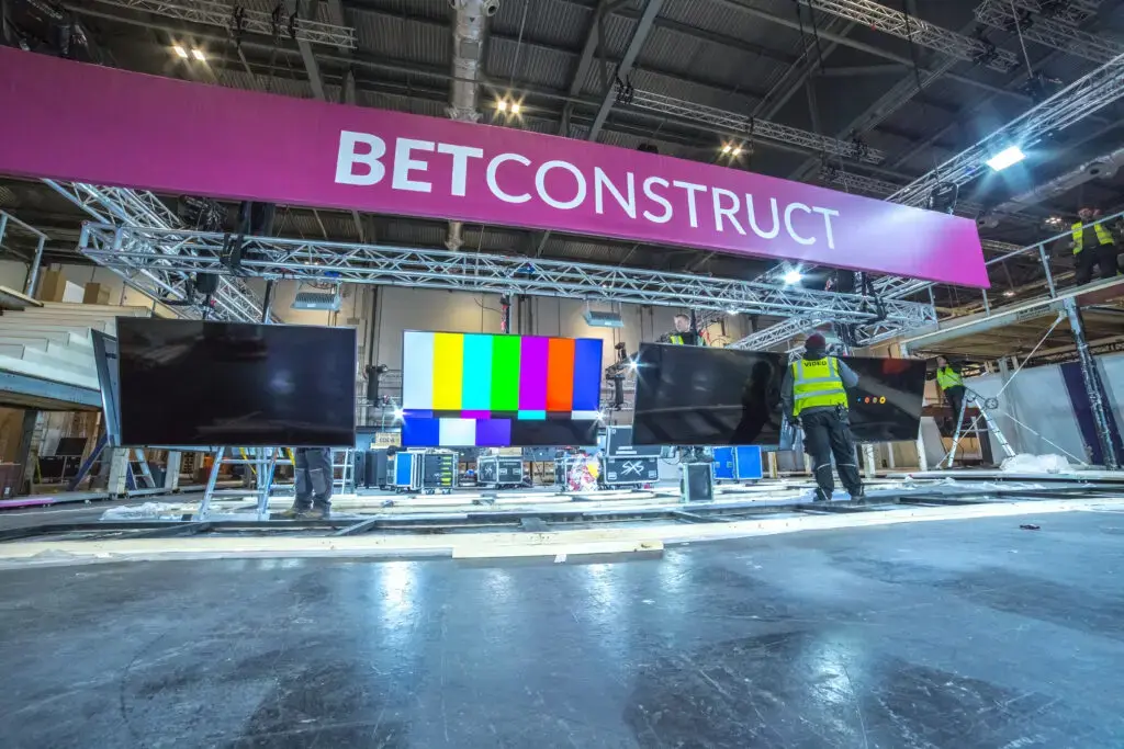 Workers setting up a BetConstruct exhibition booth with large digital screens and a purple overhead banner