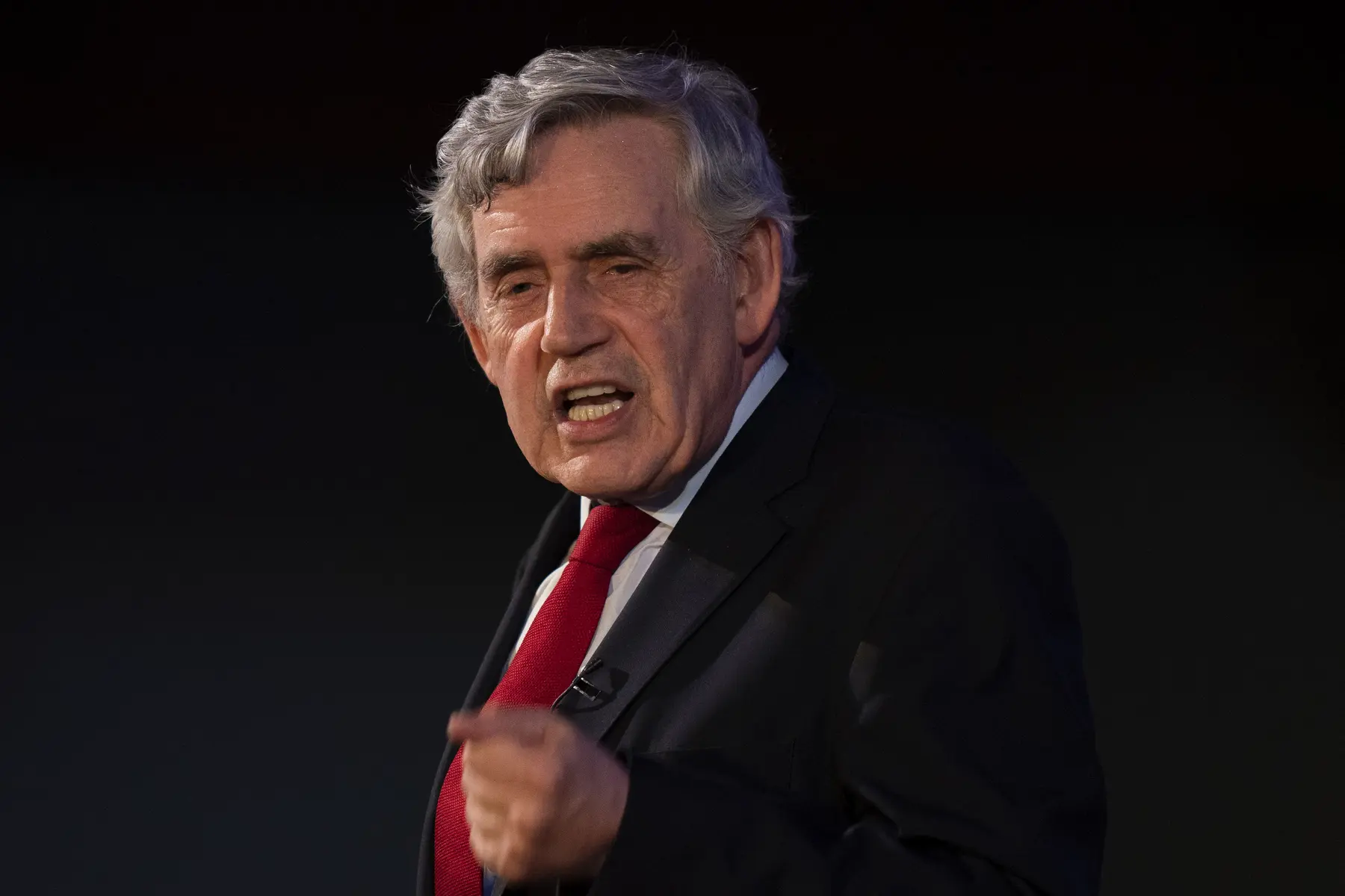 Gordon Brown speaking passionately, wearing a dark suit and red tie against a dark background.