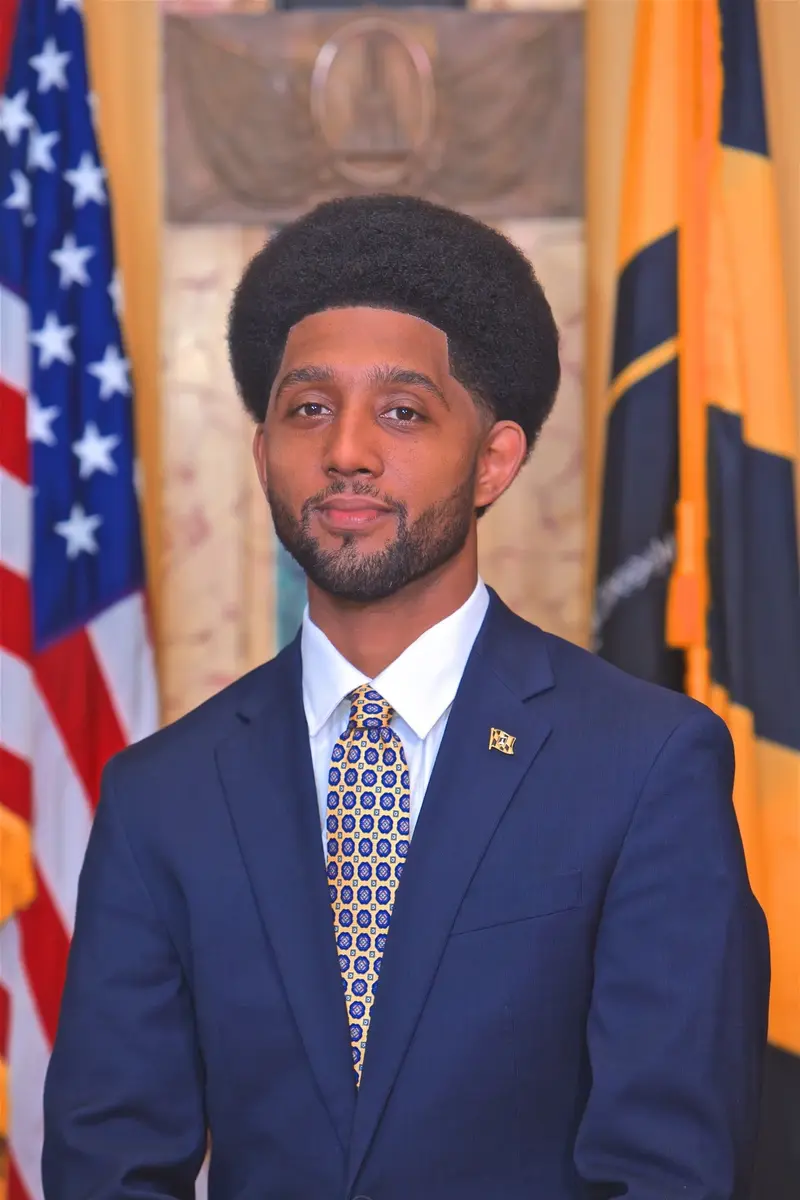 Portrait of Mayor Brandon Scott wearing a blue suit and yellow patterned tie in front of US and Maryland flags.