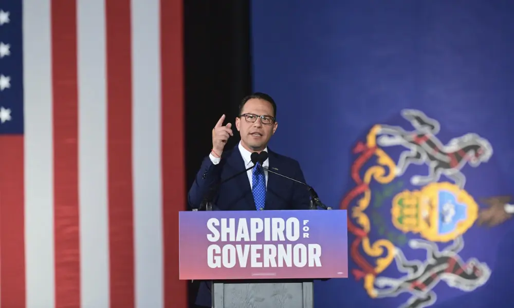 Josh Shapiro speaking at a podium with a sign reading Shapiro for Governor in front of a US flag.