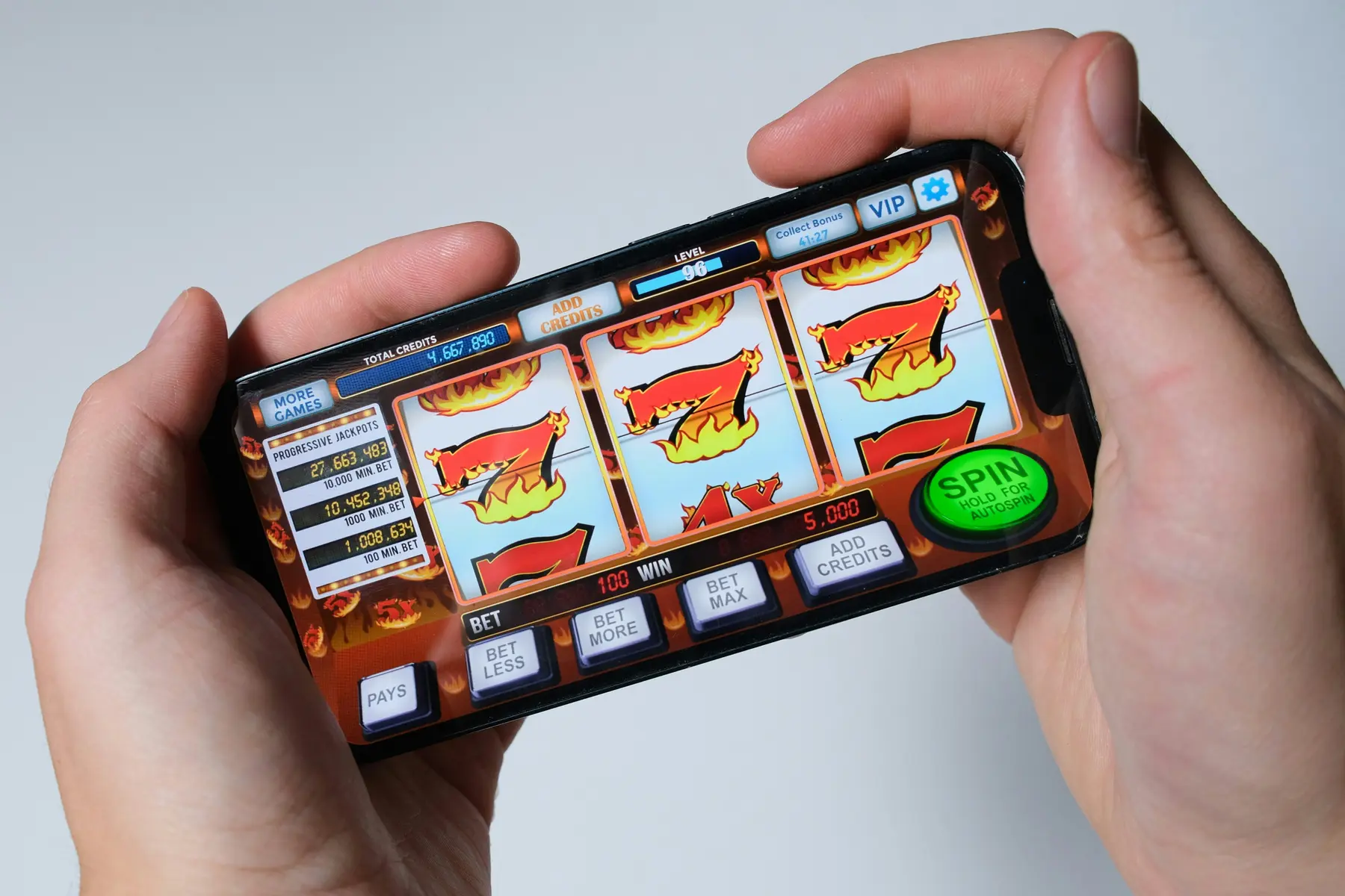 A person holding a smartphone displaying a colorful digital slot machine game with flaming sevens.