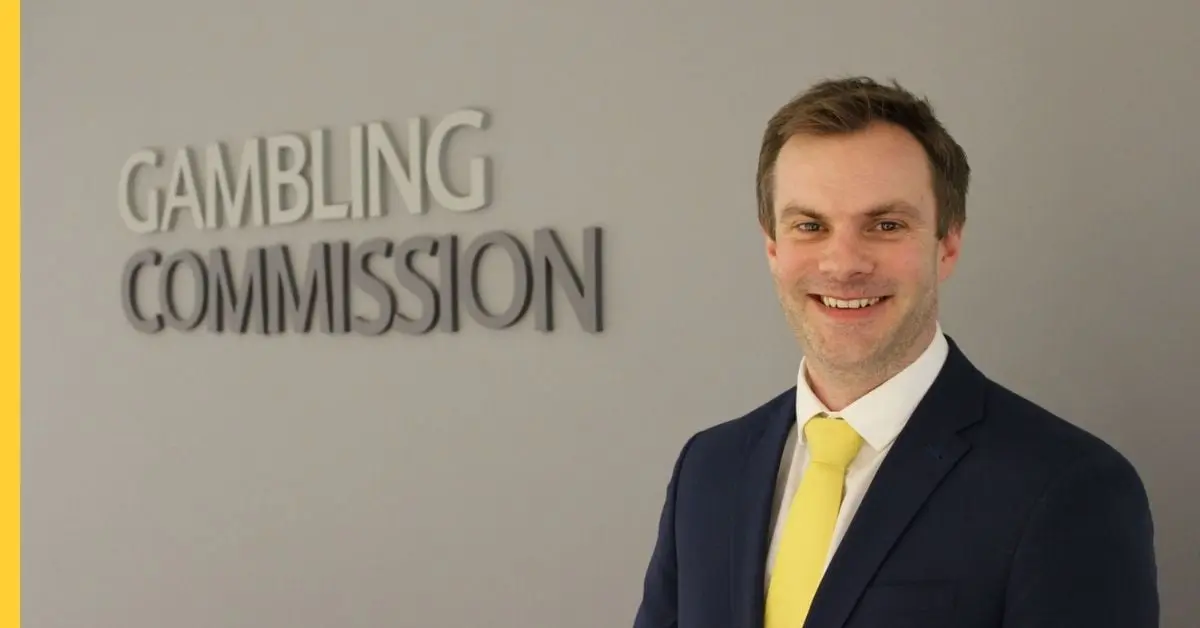 Tim Miller smiling in a navy suit and yellow tie, standing in front of a Gambling Commission wall sign.